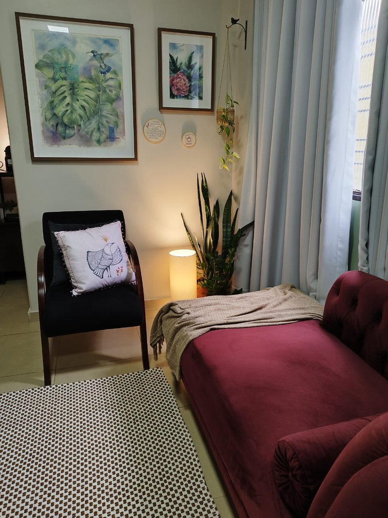 Consultório de Josiane com divã vinho, poltrona, aquarelas, plantas e luz junto à cortina.